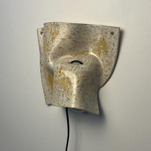 May include: A wall-mounted ceramic light fixture with an irregular, folded shape. The surface features a speckled cream and beige glaze with patches of yellow. A black power cord extends from the center, and two small holes are visible at the top corners.