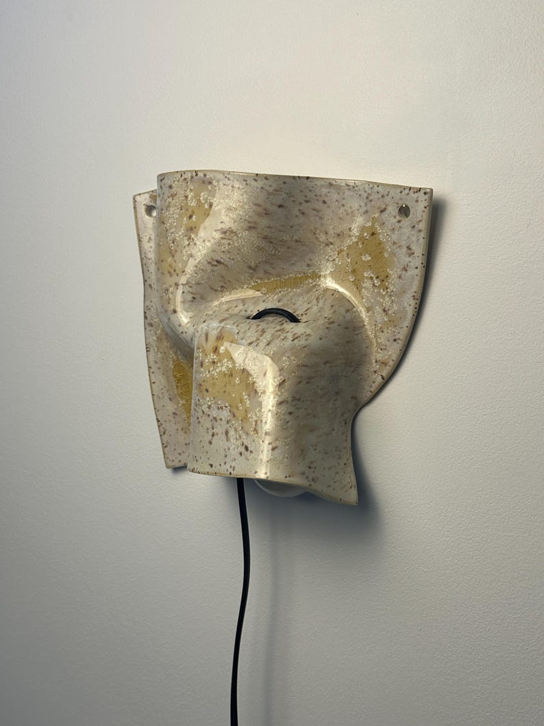 May include: A wall-mounted ceramic light fixture with an irregular, folded shape. The surface features a speckled cream and beige glaze with patches of yellow. A black power cord extends from the center, and two small holes are visible at the top corners.