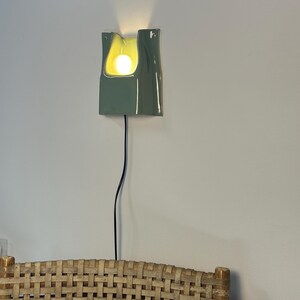 May include: A sage green ceramic wall sconce with a glossy finish and a curved, organic shape. The fixture features a central cutout housing a glowing light bulb, with a thin black power cord extending downwards from the base of the lamp.