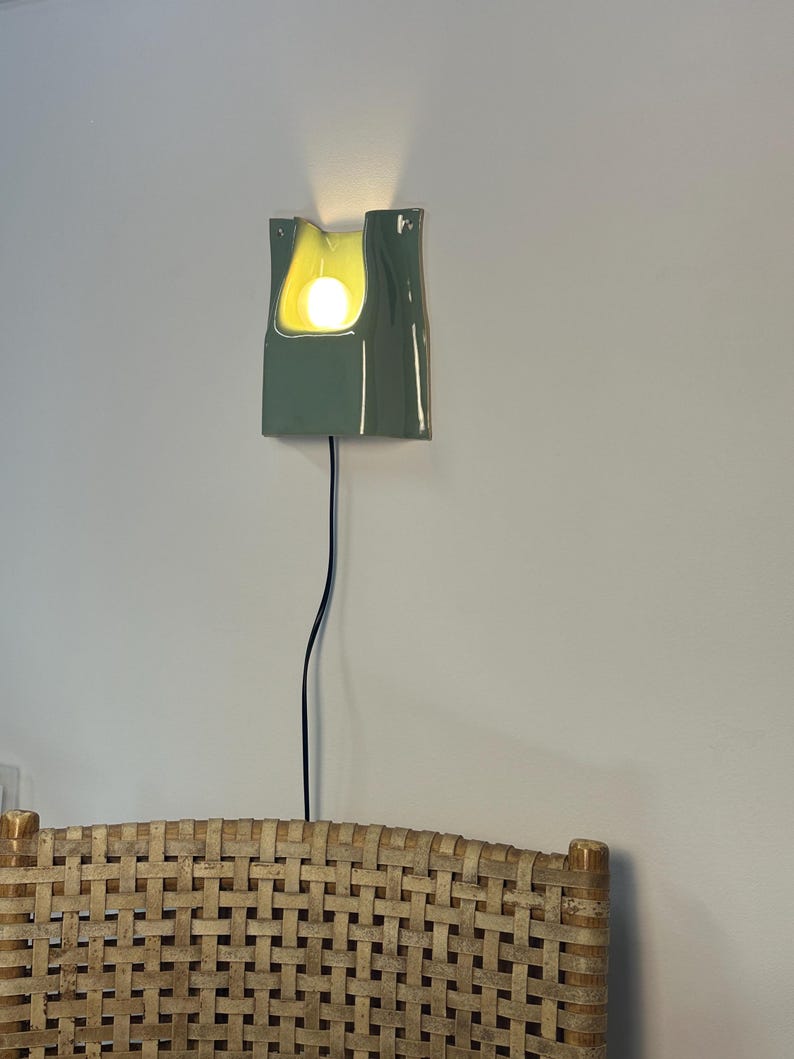 May include: A sage green ceramic wall sconce with a glossy finish and a curved, organic shape. The fixture features a central cutout housing a glowing light bulb, with a thin black power cord extending downwards from the base of the lamp.