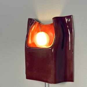 Puede incluir: Un aplique de pared de cer&aacute;mica con un acabado brillante en color burdeos intenso. La pieza presenta un centro curvo y hundido que alberga una bombilla esf&eacute;rica brillante, proyectando una luz naranja c&aacute;lida sobre la superficie interior.