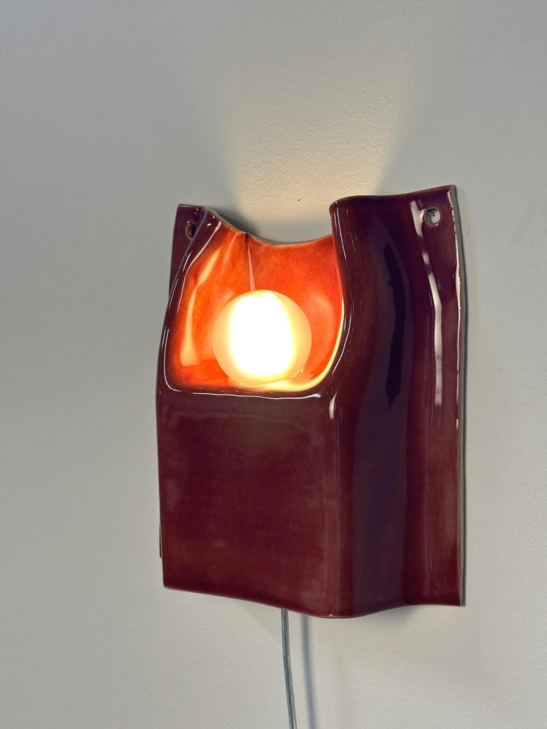 Puede incluir: Un aplique de pared de cer&aacute;mica con un acabado brillante en color burdeos intenso. La pieza presenta un centro curvo y hundido que alberga una bombilla esf&eacute;rica brillante, proyectando una luz naranja c&aacute;lida sobre la superficie interior.