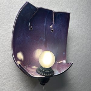 May include: A wall-mounted sconce featuring a curved, deep purple ceramic shade with a glossy finish. The fixture includes a small, round, frosted white light bulb centred at the bottom, held by a black socket with a thin, clear power cord.