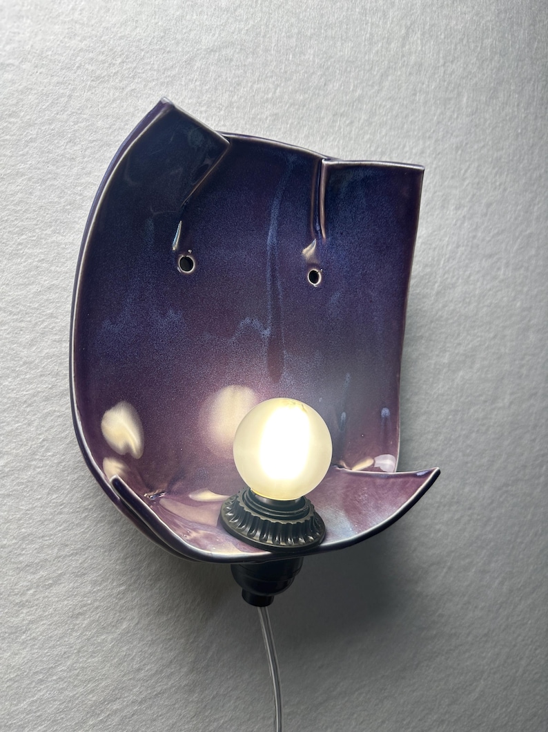 May include: A wall-mounted sconce featuring a curved, deep purple ceramic shade with a glossy finish. The fixture includes a small, round, frosted white light bulb centred at the bottom, held by a black socket with a thin, clear power cord.
