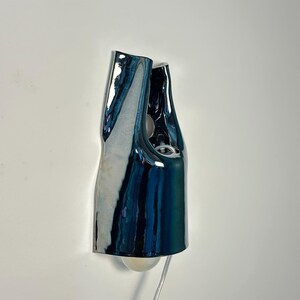 May include: A wall-mounted lamp featuring a glossy, dark blue, organic-shaped ceramic shade with a reflective surface. The fixture has an irregular, folded silhouette with a visible light bulb at the bottom and a thin, clear power cord extending downwards.