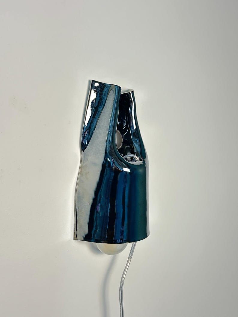 May include: A wall-mounted lamp featuring a glossy, dark blue, organic-shaped ceramic shade with a reflective surface. The fixture has an irregular, folded silhouette with a visible light bulb at the bottom and a thin, clear power cord extending downwards.