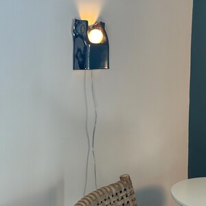 May include: A dark navy blue ceramic wall sconce with a glossy finish and an organic, irregular top edge. The fixture holds a single glowing round light bulb that casts a warm light upward and outward, connected by a thin, clear electrical cord.