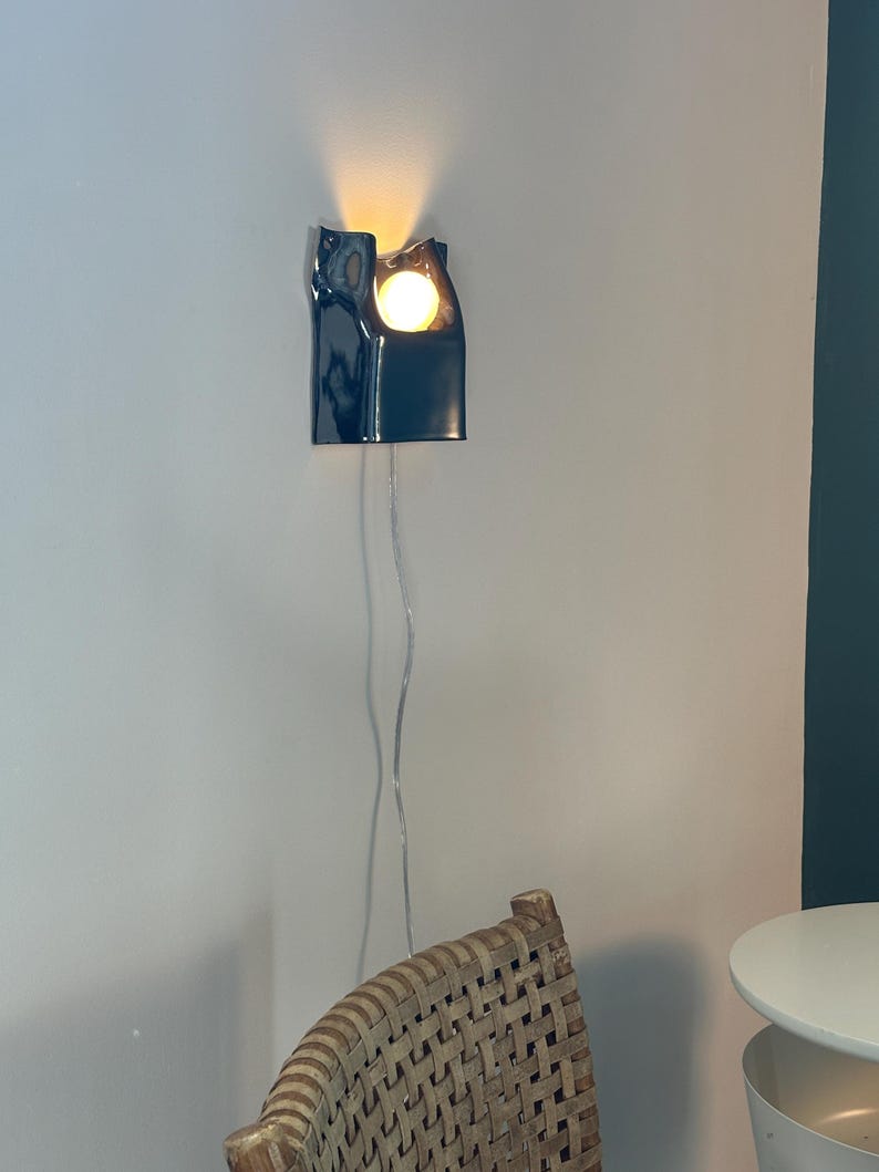 May include: A dark navy blue ceramic wall sconce with a glossy finish and an organic, irregular top edge. The fixture holds a single glowing round light bulb that casts a warm light upward and outward, connected by a thin, clear electrical cord.
