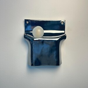 Peut inclure: Une poche murale en c&eacute;ramique bleu marine fonc&eacute; brillant avec une finition lisse et r&eacute;fl&eacute;chissante. La pi&egrave;ce rectangulaire pr&eacute;sente une section sup&eacute;rieure incurv&eacute;e et ouverte, ainsi que deux petits trous circulaires aux coins sup&eacute;rieurs pour la fixation. Une sph&egrave;re blanche est plac&eacute;e &agrave; l'int&eacute;rieur.