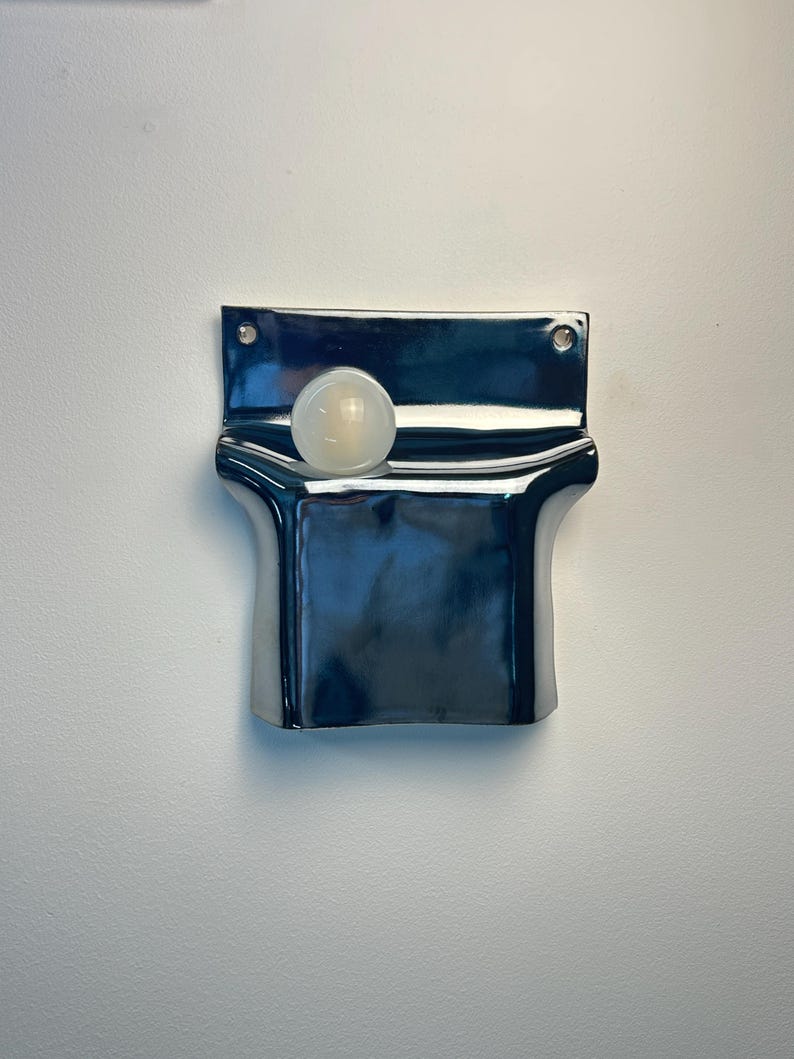 Peut inclure: Une poche murale en c&eacute;ramique bleu marine fonc&eacute; brillant avec une finition lisse et r&eacute;fl&eacute;chissante. La pi&egrave;ce rectangulaire pr&eacute;sente une section sup&eacute;rieure incurv&eacute;e et ouverte, ainsi que deux petits trous circulaires aux coins sup&eacute;rieurs pour la fixation. Une sph&egrave;re blanche est plac&eacute;e &agrave; l'int&eacute;rieur.