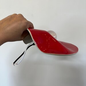 May include: A glossy red curved lamp shade with a smooth, undulating surface. A white light bulb is attached to the underside. Two exposed electrical wires, one black and one white, extend from the base of the lamp fixture.