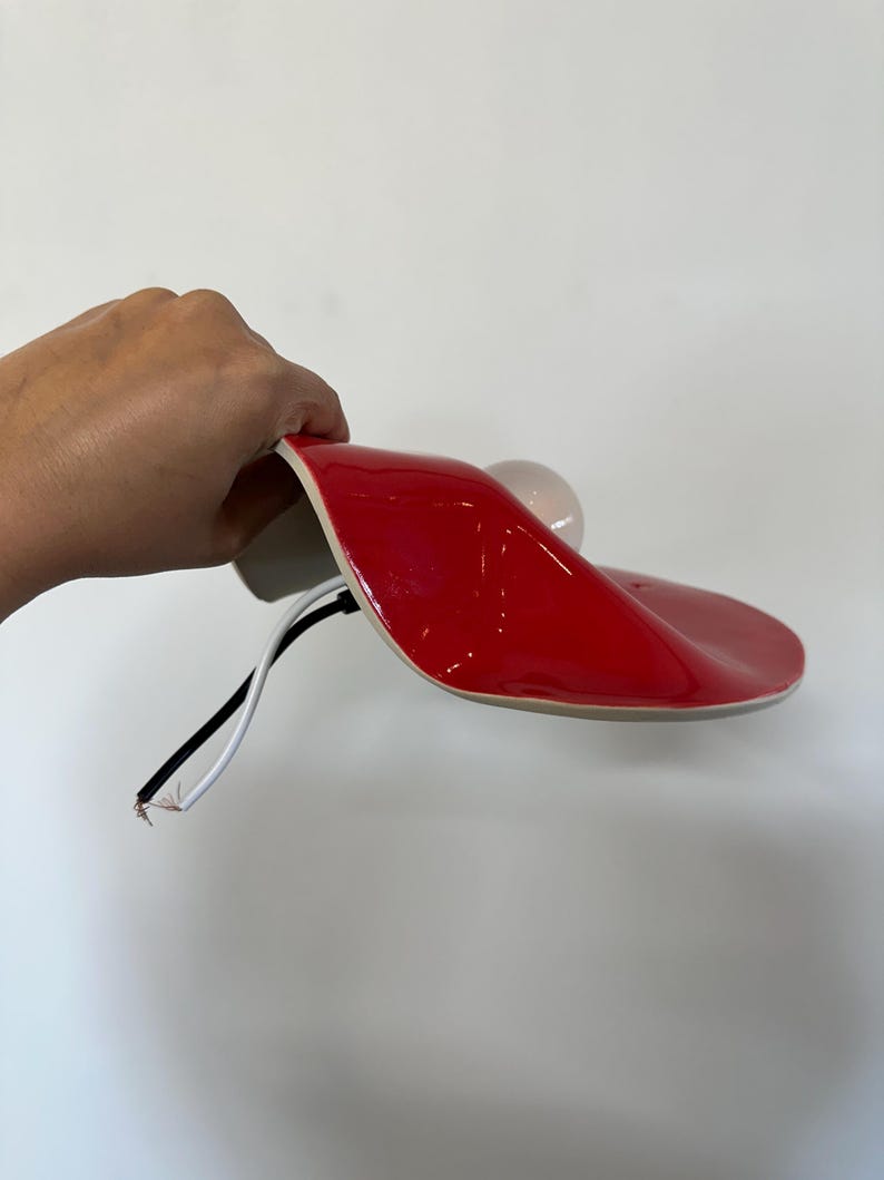 May include: A glossy red curved lamp shade with a smooth, undulating surface. A white light bulb is attached to the underside. Two exposed electrical wires, one black and one white, extend from the base of the lamp fixture.
