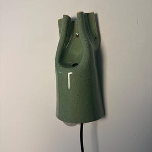 May include: A wall-mounted ceramic light fixture in a sage green color with dark speckles. The object features a vertical, elongated, and folded form with a smooth, glossy finish. A black electrical cord extends from the bottom of the fixture.