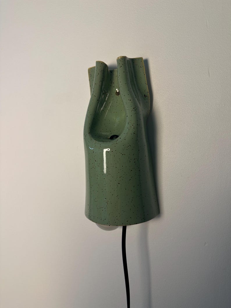 May include: A wall-mounted ceramic light fixture in a sage green color with dark speckles. The object features a vertical, elongated, and folded form with a smooth, glossy finish. A black electrical cord extends from the bottom of the fixture.