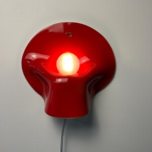May include: A glossy red wall lamp with a curved, organic shape and a central circular opening. A single round light bulb is positioned in the center, emitting a warm glow. A small mounting hole is visible at the top, and a thin cord hangs below.