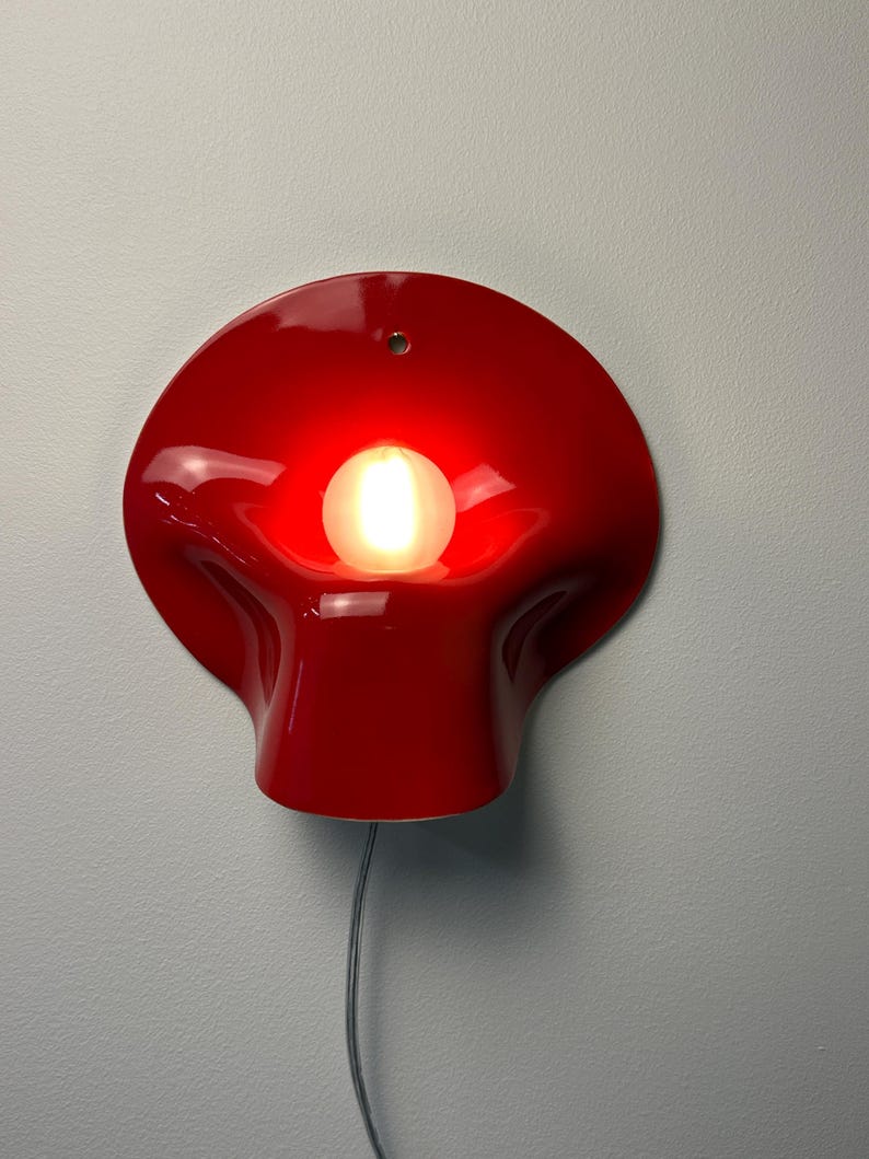 May include: A glossy red wall lamp with a curved, organic shape and a central circular opening. A single round light bulb is positioned in the center, emitting a warm glow. A small mounting hole is visible at the top, and a thin cord hangs below.