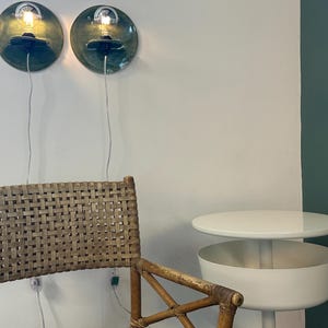 May include: Two circular wall sconces with glossy dark green ceramic backplates and exposed light bulbs, a woven rattan chair with a wooden frame, and a white two-tiered side table with a round top and a cylindrical base.