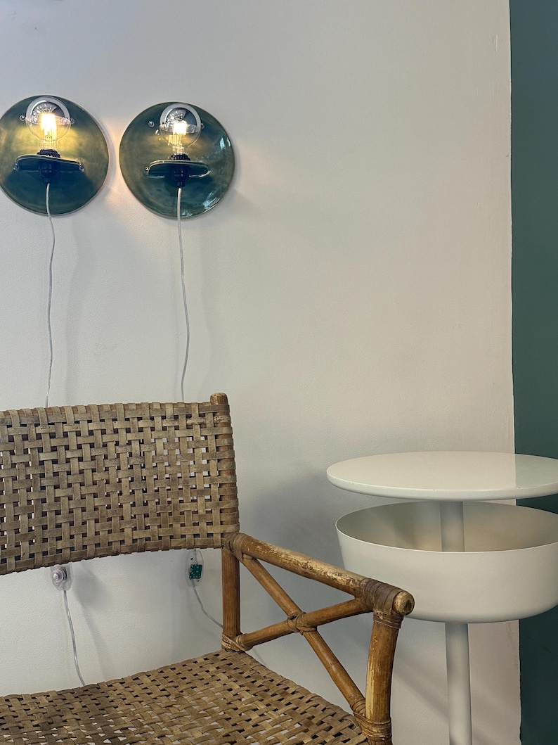 May include: Two circular wall sconces with glossy dark green ceramic backplates and exposed light bulbs, a woven rattan chair with a wooden frame, and a white two-tiered side table with a round top and a cylindrical base.