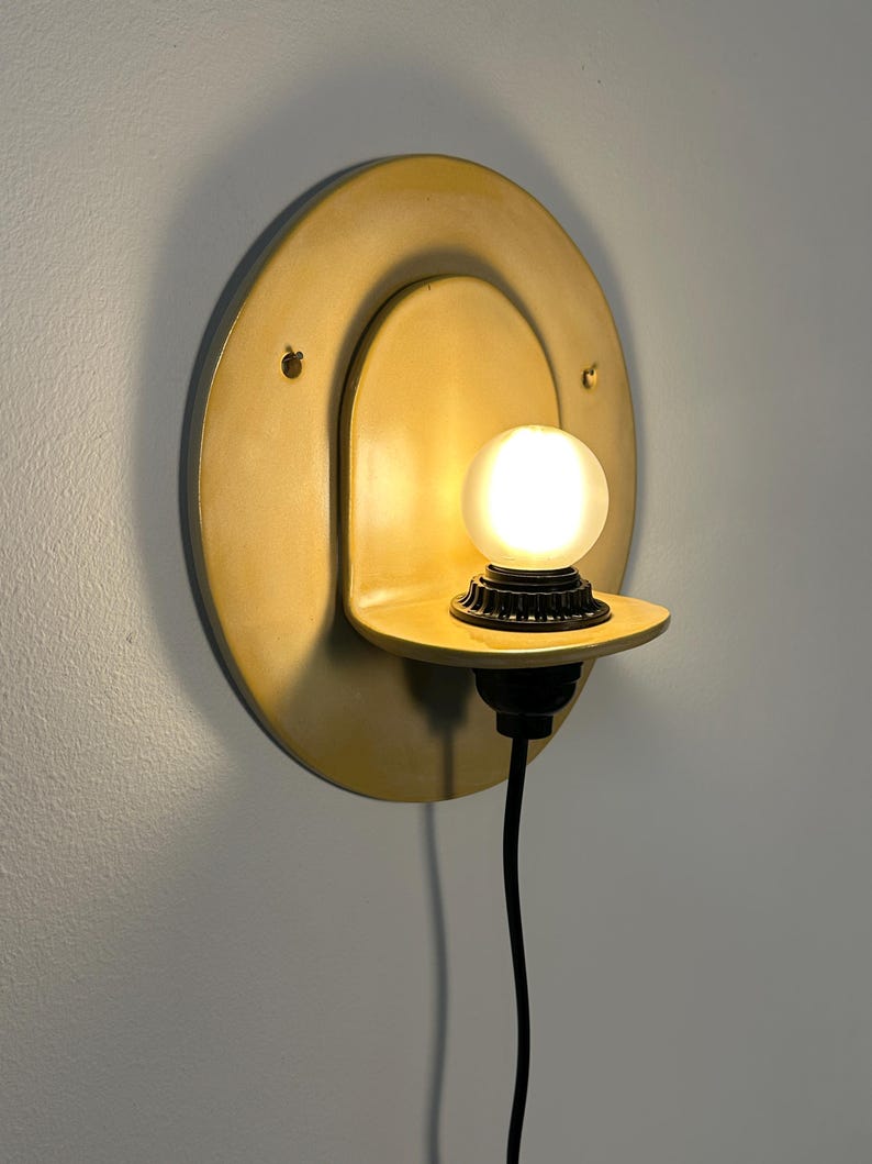 Pu&ograve; includere: Una lampada da parete con una piastra posteriore circolare gialla e un piccolo ripiano sporgente. Un portalampada nero sostiene una lampadina sferica smerigliata accesa. Un cavo di alimentazione nero pende verticalmente dalla base.