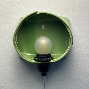 May include: A wall-mounted light fixture featuring a glossy, sage green ceramic shade with an organic, folded shape. A black socket holds a round, frosted white light bulb in the center, with a thin, clear power cord extending downward.