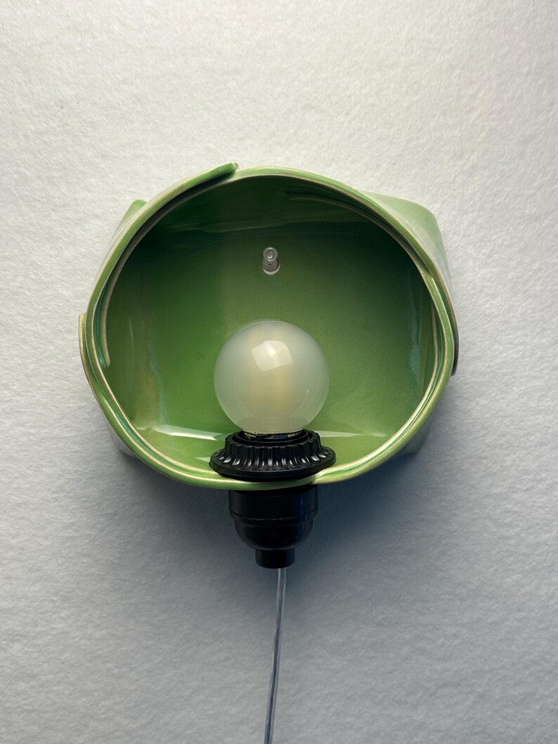 May include: A wall-mounted light fixture featuring a glossy, sage green ceramic shade with an organic, folded shape. A black socket holds a round, frosted white light bulb in the center, with a thin, clear power cord extending downward.