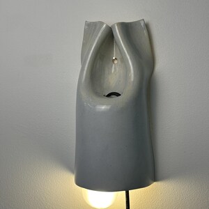 May include: A wall-mounted light fixture made of smooth, light gray ceramic with a folded, organic shape. The fixture features a central opening with a visible black cord and a glowing light bulb extending from the bottom edge of the ceramic body.