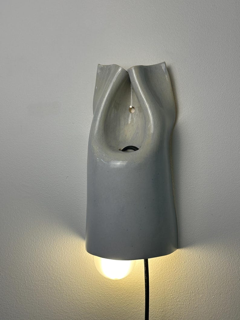 May include: A wall-mounted light fixture made of smooth, light gray ceramic with a folded, organic shape. The fixture features a central opening with a visible black cord and a glowing light bulb extending from the bottom edge of the ceramic body.