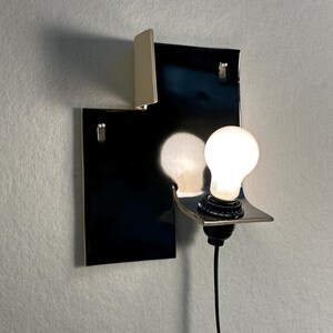 Pode incluir: Uma arandela de parede moderna com uma base retangular preta e um elemento branco. Uma placa de metal prateado suporta uma l&acirc;mpada redonda acesa. A arandela &eacute; montada em uma parede cinza claro.