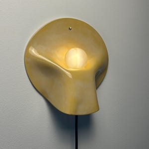 May include: A wall-mounted lamp featuring a smooth, pale yellow ceramic shade with a curved, organic shape. A single, round, frosted glass bulb sits in the center of the shade, emitting a soft glow. A black power cord extends from the bottom.