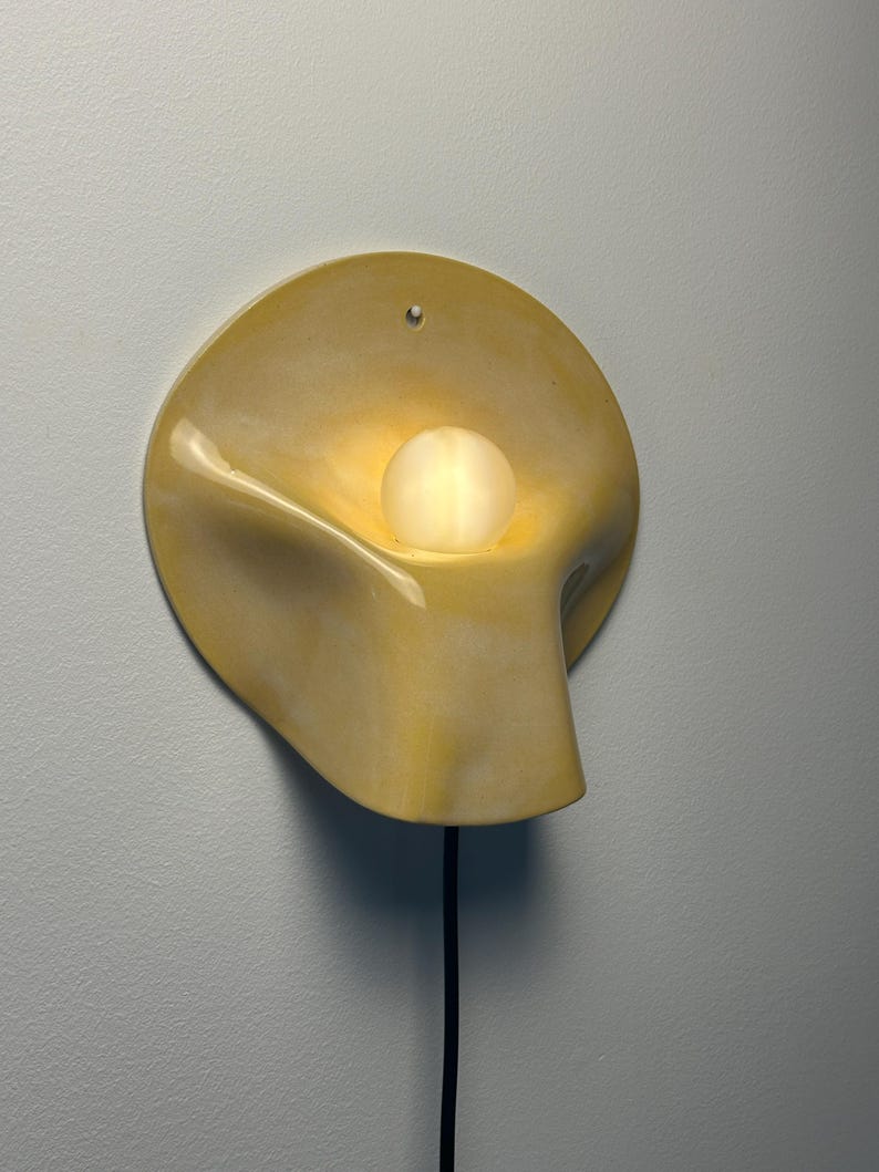May include: A wall-mounted lamp featuring a smooth, pale yellow ceramic shade with a curved, organic shape. A single, round, frosted glass bulb sits in the center of the shade, emitting a soft glow. A black power cord extends from the bottom.