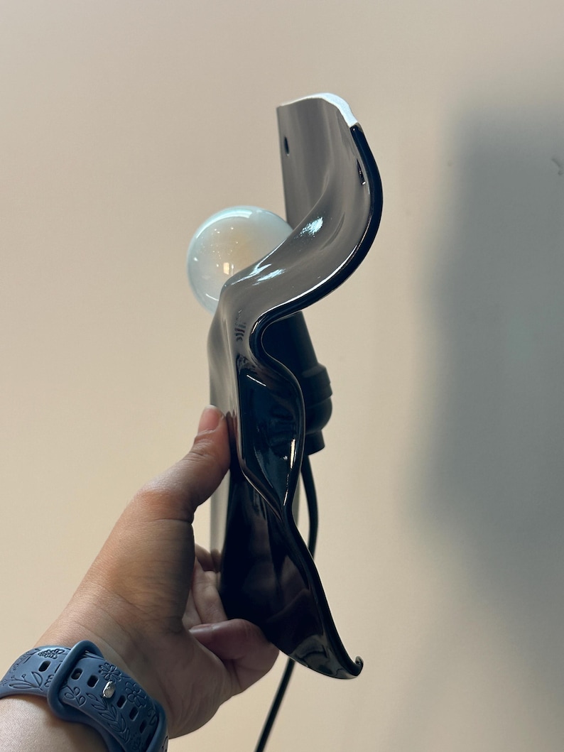 Puede incluir: Una l&aacute;mpara de pared con una pantalla de vidrio negro brillante, ondulada y de forma org&aacute;nica. El accesorio incluye una bombilla redonda de color blanco esmerilado colocada en el centro. La estructura tiene una superficie lisa, reflectante y curva.