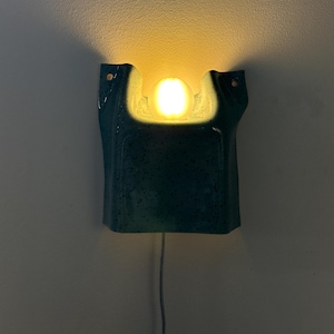 May include: A wall-mounted sconce featuring a teal ceramic housing with a speckled finish and a central cutout. A single glowing, warm-toned spherical light bulb is positioned within the cutout, casting a soft light upward against the wall.