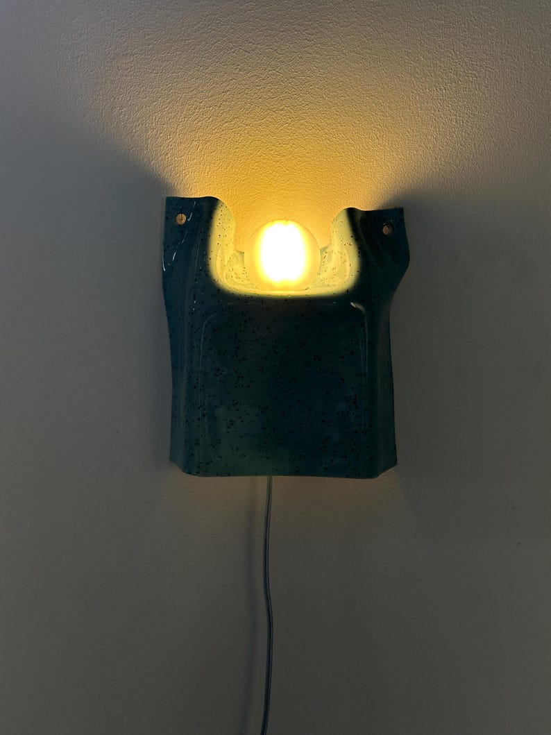 May include: A wall-mounted sconce featuring a teal ceramic housing with a speckled finish and a central cutout. A single glowing, warm-toned spherical light bulb is positioned within the cutout, casting a soft light upward against the wall.