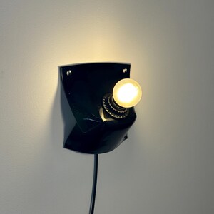 May include: A black wall-mounted lamp featuring a curved, shield-like base with two visible mounting holes at the top corners. A single round, glowing light bulb is centered in a metallic socket, and a black power cord extends downward from the base.