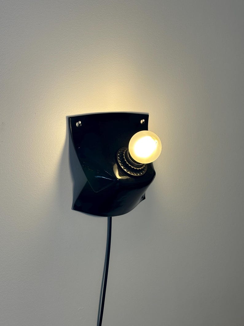 May include: A black wall-mounted lamp featuring a curved, shield-like base with two visible mounting holes at the top corners. A single round, glowing light bulb is centered in a metallic socket, and a black power cord extends downward from the base.