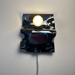 May include: A wall-mounted lamp featuring a glossy, dark, wavy ceramic backplate with a central circular socket holding a single glowing, frosted spherical light bulb. A thin, light-colored electrical cord extends downward from the base.
