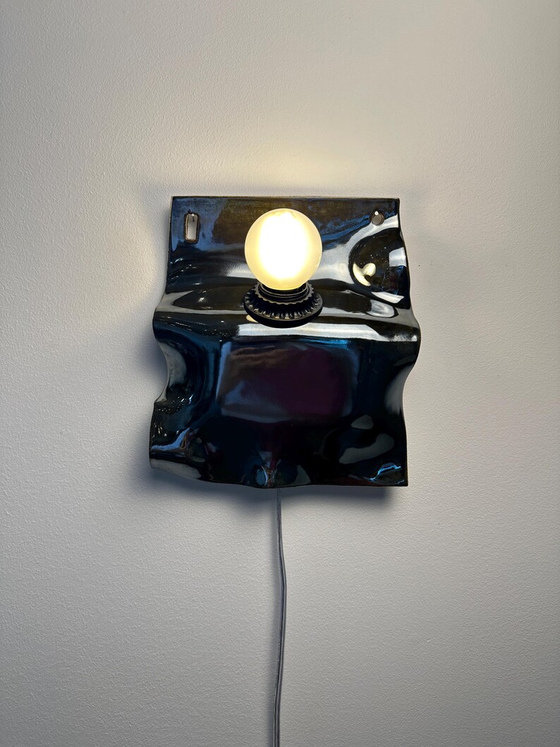 May include: A wall-mounted lamp featuring a glossy, dark, wavy ceramic backplate with a central circular socket holding a single glowing, frosted spherical light bulb. A thin, light-colored electrical cord extends downward from the base.