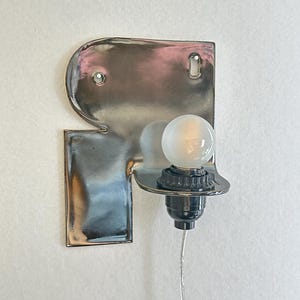 May include: A wall-mounted lamp featuring a reflective, silver-toned metallic backplate with a curved, protruding shelf. A black socket holds a small, frosted spherical light bulb, and a thin, clear electrical cord extends downward from the base.