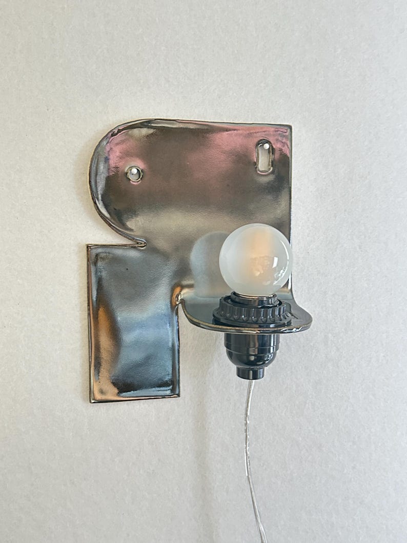 May include: A wall-mounted lamp featuring a reflective, silver-toned metallic backplate with a curved, protruding shelf. A black socket holds a small, frosted spherical light bulb, and a thin, clear electrical cord extends downward from the base.