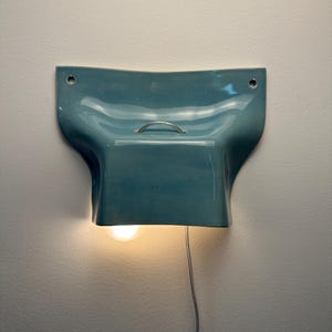 K&ouml;nnte beinhalten: Eine Wandleuchte mit einem gl&auml;nzenden, blaugr&uuml;nen Keramikschirm in geschwungener, organischer Form. Die Leuchte verf&uuml;gt &uuml;ber einen kleinen silbernen Metallgriff an der Oberseite, zwei Befestigungsl&ouml;cher an den oberen Ecken und eine leuchtende Gl&uuml;hbirne.