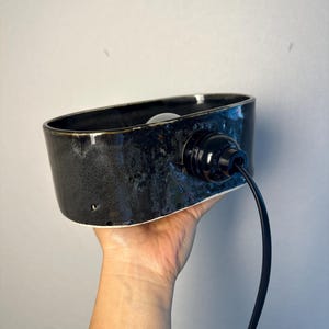 May include: A black ceramic wall sconce with a glossy finish. The sconce has an oval shape and a black electrical cord. The light fixture is held by a hand against a grey wall.
