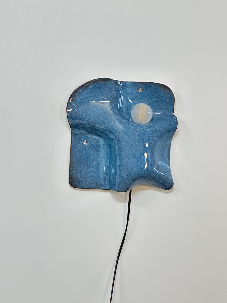 May include: A wall-mounted ceramic light fixture with a glossy blue glaze and an irregular, organic shape. The piece features a central circular white light bulb and a black power cord extending from the bottom edge of the textured blue surface.