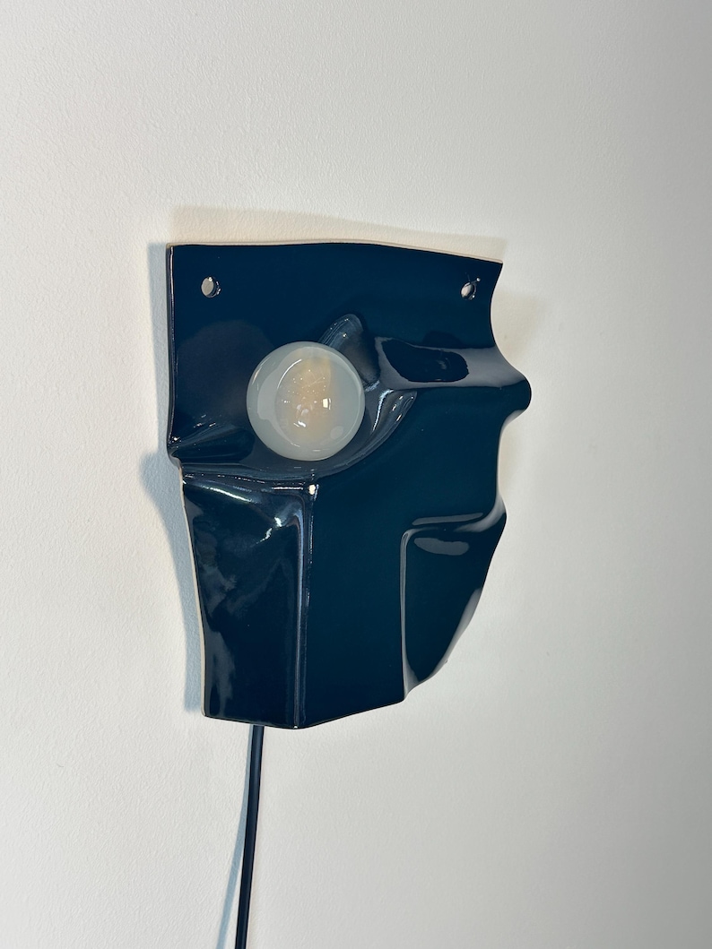 Puede incluir: Una l&aacute;mpara de pared con una base de cer&aacute;mica azul marino oscuro brillante de forma irregular y plegada. Una bombilla redonda de color blanco esmerilado est&aacute; centrada en una abertura circular. Un cable de alimentaci&oacute;n negro sale por debajo.