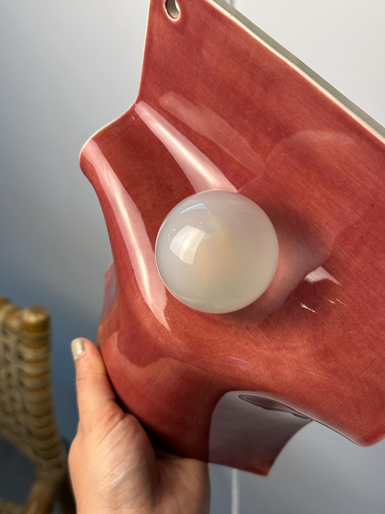 May include: A wall-mounted lamp featuring a glossy, dusty rose-colored ceramic shade with a wavy, organic shape. A single, spherical, frosted white glass light bulb is centered on the front of the ceramic fixture, which includes a small hole at the top.