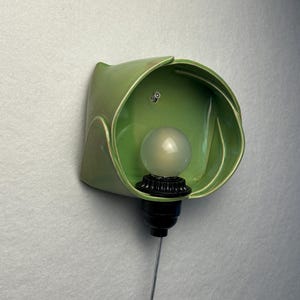 May include: A wall-mounted light fixture featuring a glossy, sage green ceramic shade with a curved, petal-like form. The fixture includes a black socket holding a round, frosted white light bulb, with a thin silver cord extending downward.