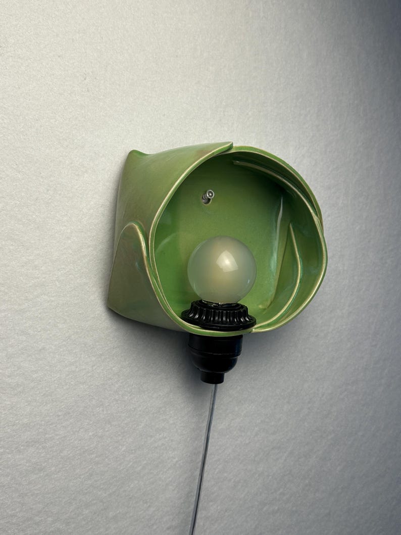 May include: A wall-mounted light fixture featuring a glossy, sage green ceramic shade with a curved, petal-like form. The fixture includes a black socket holding a round, frosted white light bulb, with a thin silver cord extending downward.