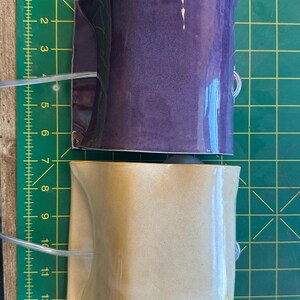 May include: Two curved ceramic wall pockets, one in a glossy deep purple and one in a glossy tan finish. Each piece features a clear plastic tube attached to the side. They are positioned on a green cutting mat with yellow grid lines measuring 13 inches.