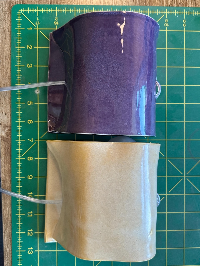 May include: Two curved ceramic wall pockets, one in a glossy deep purple and one in a glossy tan finish. Each piece features a clear plastic tube attached to the side. They are positioned on a green cutting mat with yellow grid lines measuring 13 inches.