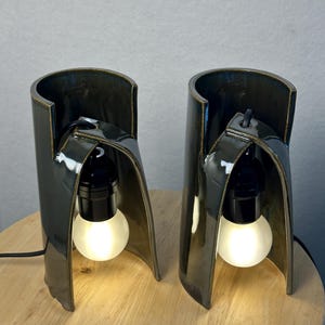 Puede incluir: Un par de l&aacute;mparas de mesa de cer&aacute;mica en color verde oliva oscuro con una estructura cil&iacute;ndrica curva. Cada l&aacute;mpara cuenta con un casquillo negro que sostiene una bombilla redonda de vidrio esmerilado blanco con luz c&aacute;lida y cable.