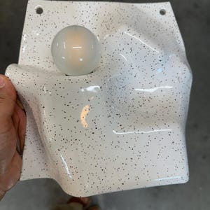 May include: A white wall sconce with a speckled black pattern and a glossy finish. The fixture features an irregular, undulating surface with a single round, glowing light bulb integrated into the left side and two mounting holes at the top corners.